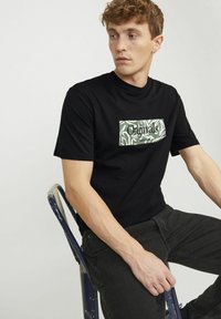 Schwarzes T-Shirt mit kurzen Ärmeln, das ein rechteckiges grün-weißes Grafikdesign mit dem schwarzen Text "Originals" zeigt. Der Träger sitzt auf einem Stuhl und trägt dunkle Jeans.