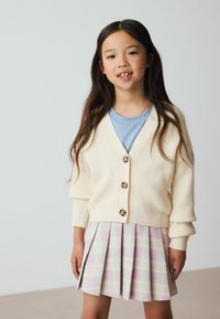 Cardigan côtelé crème avec des boutons dorés, associé à une jupe plissée à carreaux pastel dans des tons de rose, jaune et violet. T-shirt bleu clair en dessous.