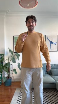 Maglietta a maniche lunghe a coste giallo senape con scollatura a girocollo, abbinata a pantaloni cargo beige chiaro. Sullo sfondo si vedono una pianta verde e un'opera d'arte incorniciata.