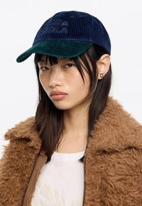 Cappello in corduroy blu con visiera in corduroy verde, caratterizzato da un logo embossato sulla parte anteriore. Chi lo indossa ha i capelli lisci ed è vestito con una giacca marrone in peluche.