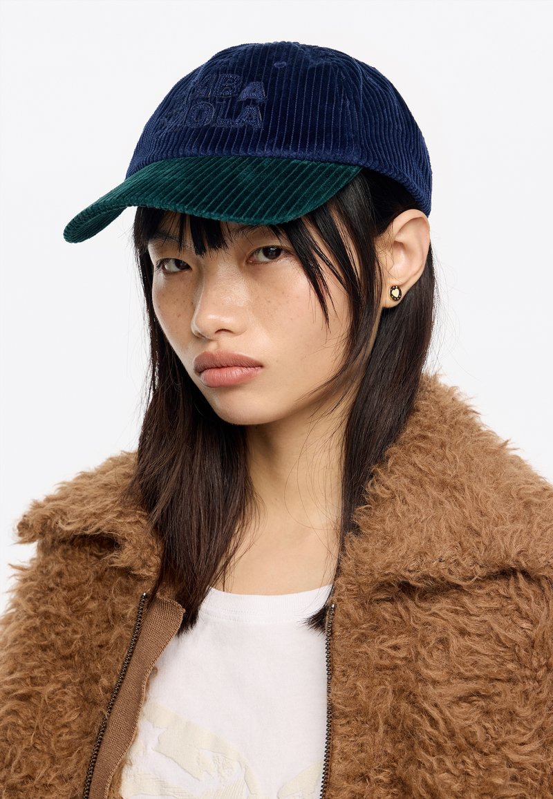 Cappello in corduroy blu con visiera in corduroy verde, caratterizzato da un logo embossato sulla parte anteriore. Chi lo indossa ha i capelli lisci ed è vestito con una giacca marrone in peluche.