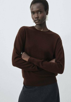 Brown sweater, round neck, long sleeves, soft texture. Model has short hair with large clear earrings, arms crossed in front. Dark background.