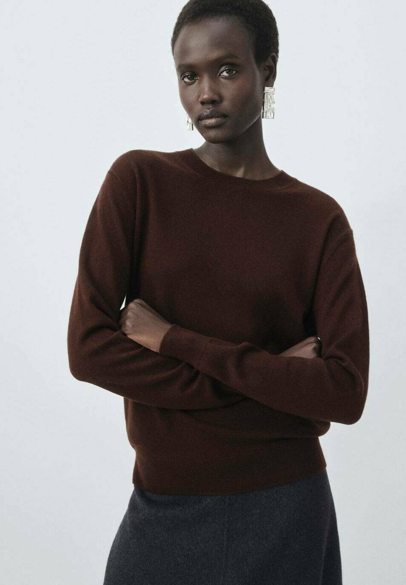 Pull marron, col rond, manches longues, texture douce. Le modèle a les cheveux courts et de grandes boucles d'oreilles transparentes, bras croisés devant lui. Fond sombre.
