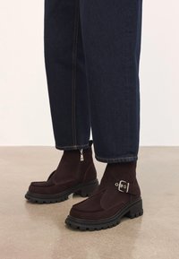 Botines de ante marrón oscuro con suelas gruesas negras, hebilla de metal y cremallera lateral, llevados con jeans azul oscuro sobre un suelo pulido.