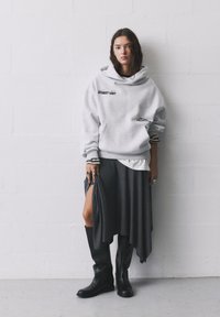 Sivý oversized sveter s potlačou "STREET UNIT", vrstvený nad sivou asymetrickou sukňou. Čierne čižmy po kolená dopĺňajú vzhľad.