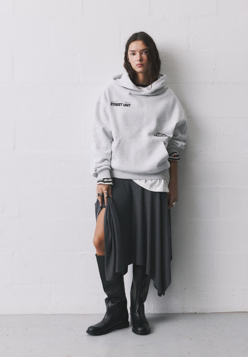 Sivý oversized sveter s potlačou "STREET UNIT", vrstvený nad sivou asymetrickou sukňou. Čierne čižmy po kolená dopĺňajú vzhľad.