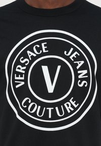 Svart bomull T-skjorte med en fremtredende hvit sirkulær logo med "VERSACE JEANS COUTURE" og et sentralt stilisert "V."