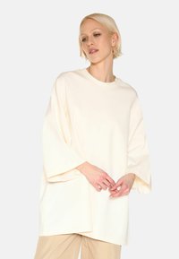 Krémové oversized tričko se širokými rukávy, kulatým výstřihem a hladkým povrchem, kombinované s světle béžovými kalhotami.