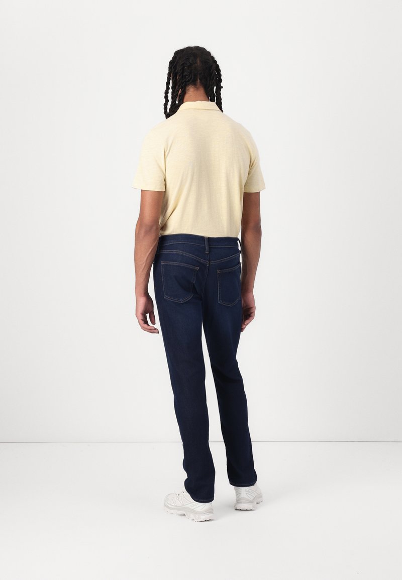 Camisa de manga corta amarilla clara, vaqueros de mezclilla azul oscuro y zapatillas blancas. La camisa tiene un acabado texturizado; los vaqueros cuentan con bolsillos traseros clásicos.