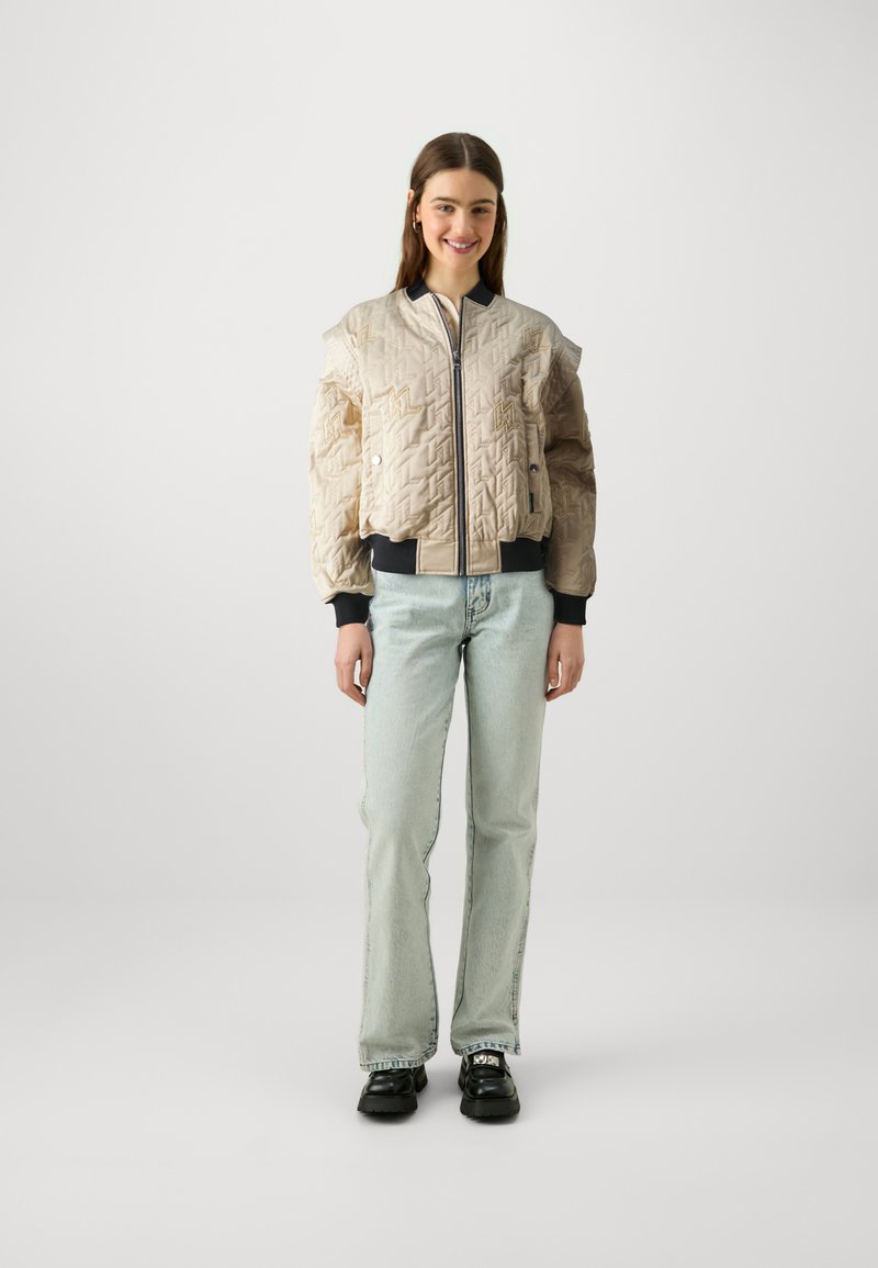 Beige gesteppte Bomberjacke mit gepr⊘gten geometrischen Mustern, gerippten Bündchen und einem Reißverschluss. Kombiniert mit hellblauer Denim und schwarzen Schuhen.