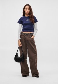 Tumši zils cropped T-krekls ar baltu grafiku, valkāts virs gaiši pelēkas garām piedurknēm krekla. Brūnas plaša piegriezuma bikses, melns jostas siksna un melna soma.