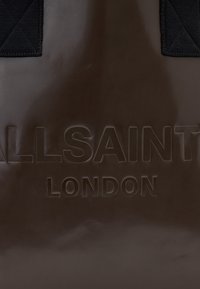 Borsa tote marrone lucida con logo in rilievo "ALLSAINTS LONDON", con accenti di tessuto trapuntato nero nella parte superiore. Texture complessivamente liscia.