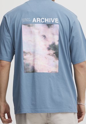 Person, der ein hellblaues Hemd mit einem wolkenartigen Muster und dem Text "ARCHIV" auf dem Rücken trägt, steht mit entspannten Armen an der Seite.