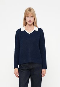 Pulover tricotat de culoare albastru marin, cu decolteu în V, având o cămașă albă cu guler dedesubt. Mâneci lungi, textură moale și o siluetă în formă de A.