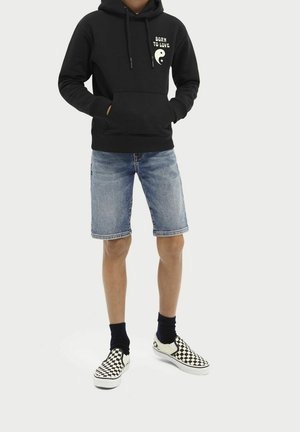 Un hoodie noir avec le texte "NÉ POUR AIMER" et un graphique yin-yang. Associé à un short en jean bleu et des chaussures à enfiler à carreaux.