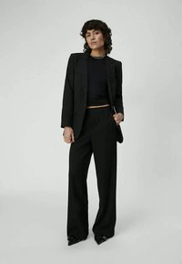 Donna con i capelli ricci che indossa un blazer nero, una maglietta nera, pantaloni larghi neri e scarpe nere a punta, posando su uno sfondo leggero e neutro.