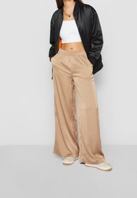 Veste bomber en satin noir, top court blanc et pantalon en satin beige à jambes larges. Les chaussures sont beiges avec des accents blancs et orange ; texture lisse.