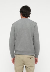 Mees, kellel on seljas lihtne hall pikasleeve sweatshirt ja beežid püksid, seisab kaamera poole seljaga valge tausta ees.