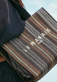 Woven tote bag in brown and black stripes, featuring a white logo "LE MARAIS PARIS." Textured surface with contrasting pattern details.