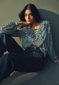 Blouse transparente avec un col froncé et un motif tribal vert-bleu, associée à un jean en denim foncé taille haute et une ceinture noire.