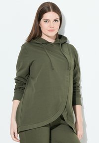 Mujer de piel clara y cabello castaño lleva una camiseta de manga larga con capucha de color verde oliva y pantalones a juego contra un fondo blanco.