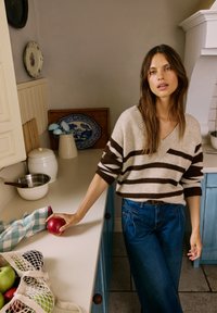 Bela in rjava črtasta pulover s špicastim izrezom, v kombinaciji z modrimi visokim pasom jeans hlačami. Rdeče jabolko v roki, kuhinjski ambient z lesenimi dodatki.