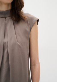 Blusa a maniche corte e collo alto in un delicato taupe chiaro. Tessuto liscio con pieghe verticali sul davanti, leggera lucentezza e vestibilità su misura.