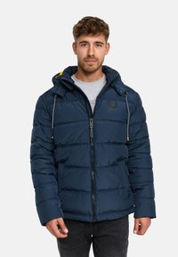 Giacca imbottita navy con cappuccio, caratterizzata da segmenti trapuntati, tasselli grigi e una toppa nera con logo sul petto. Chiusura frontale con zip e tasche.