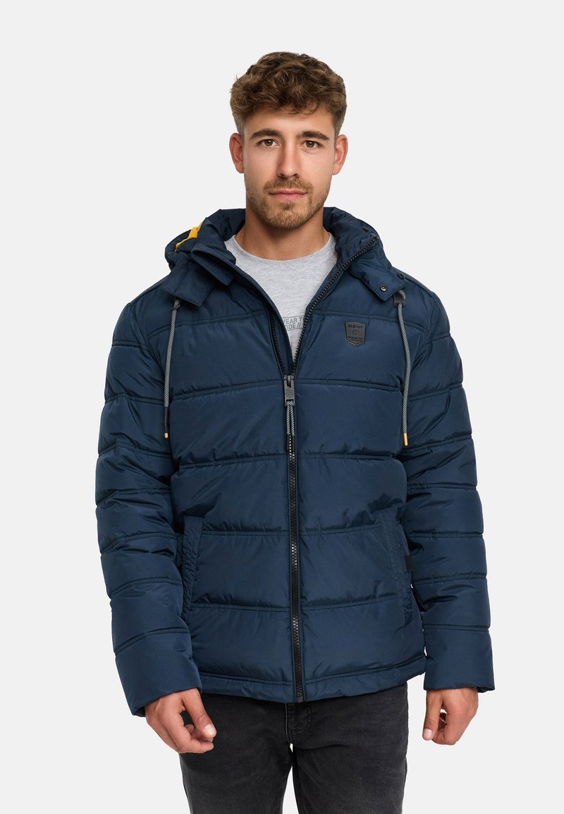 Giacca imbottita navy con cappuccio, caratterizzata da segmenti trapuntati, tasselli grigi e una toppa nera con logo sul petto. Chiusura frontale con zip e tasche.