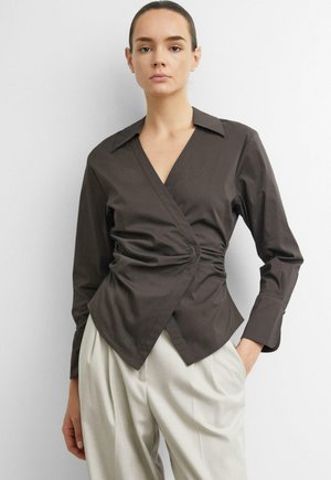 Frau trägt eine dunkel gehaltene Wickelbluse mit Kragen und langen Ärmeln, kombiniert mit hellen, hoch sitzenden Hosen, steht vor einer schlichten Wand.