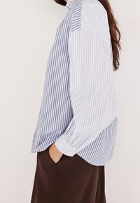 Femme portant un chemisier à manches longues rayé bleu et blanc avec un col à volants et une jupe marron foncé, main dans la poche, posture de profil.