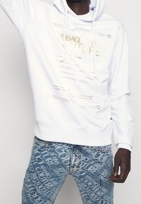 Vit hoodie med guld- och silverfärgad grafisk text, dragsko, kombinerad med blå jeans med tryck i helmönster som har en urblekt design.