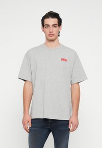 Sivé bavlnené tričko s krátkymi rukávmi, voľným strihom a červeným logom "DIESEL" na ľavej strane hrudníka. Jednoduchý dizajn a čisté línie.