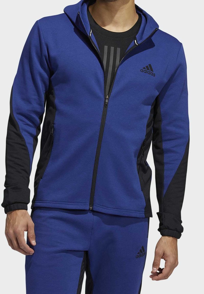 Sweat à capuche zippé bleu avec accents noirs, texture côtelée et poignets ajustés. Présente un col montant et le logo adidas sur la poitrine.