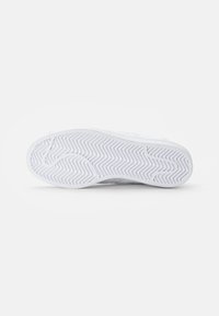 White sneaker with a textured rubber sole featuring a zigzag pattern. Upper made of smooth material with minimal branding. Clean and simple design.