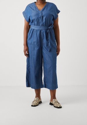 Personne portant une combinaison courte en denim bleu avec une ceinture nouée à la taille et des mocassins à motif peau de serpent sur un fond uni.
