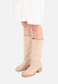 Μπεζ δερμάτινες μπότες μέχρι το γόνατο με λεία υφή, στρογγυλή μύτη και χαμηλό τετράγωνο τακούνι. Περιλαμβάνει διακοσμητική ραφή στην κνήμη.