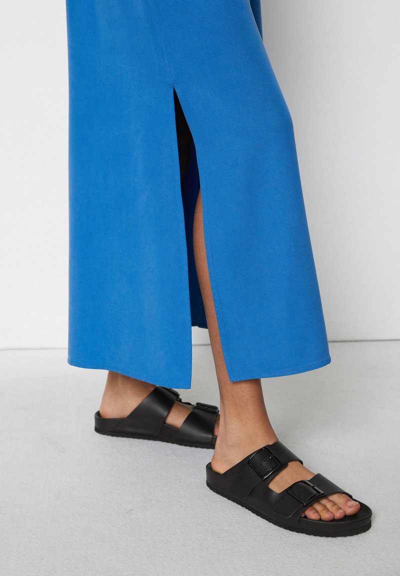 Blaue, bodenlange Bluse mit seitlichen Schlitzen, aus glattem Stoff. Schwarze Riemensandalen mit zwei verstellbaren Schnallen und strukturierten Sohlen.