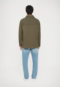 Giacca in corduroy verde oliva con colletto e maniche lunghe, abbinata a pantaloni in denim azzurro chiaro. La giacca ha una vestibilità dritta e una texture sottile.