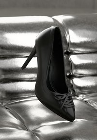 Black high-heeled pump with pointed toe and bow on shiny silver tufted leather surface.
