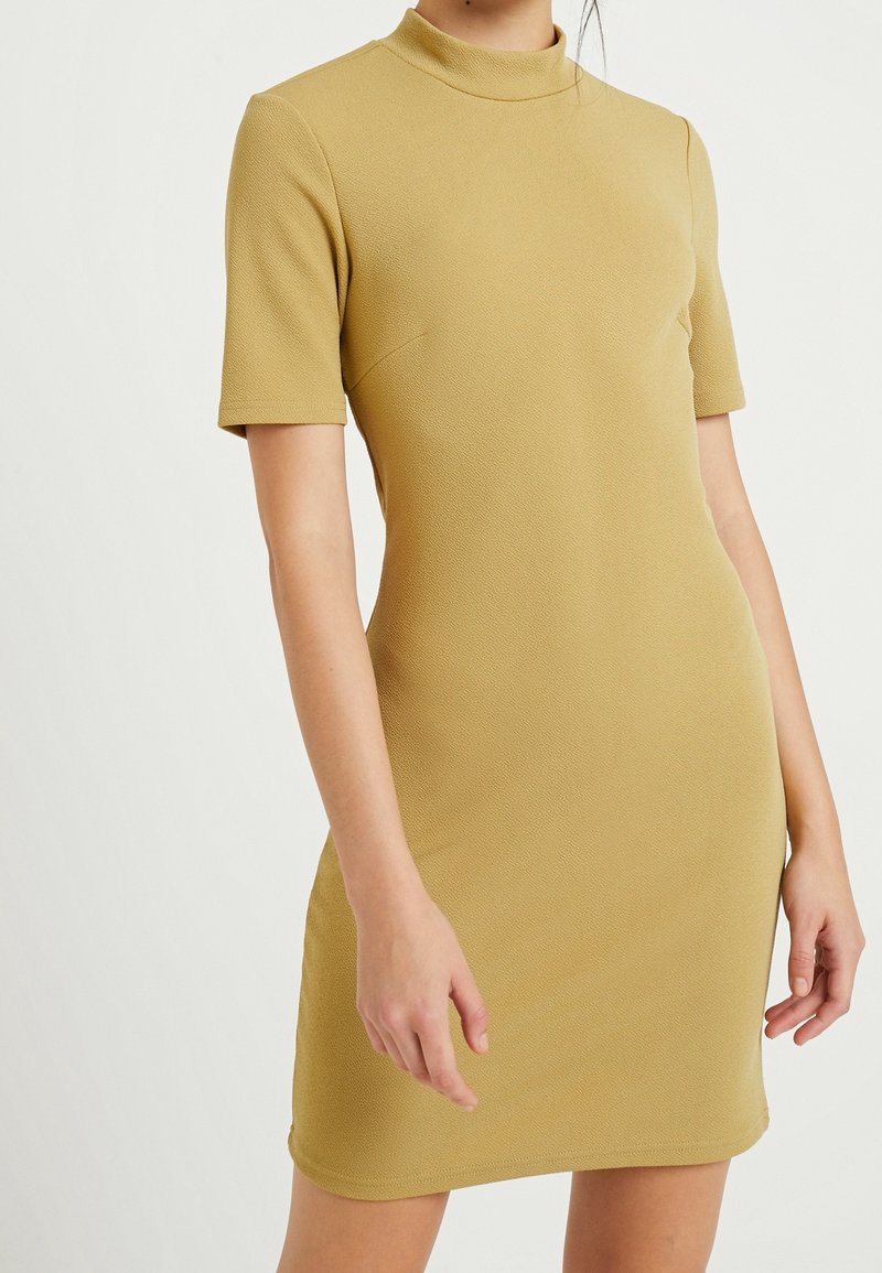 Robe beige à manches courtes ajustée avec un col rond haut, en tissu texturé, légèrement évasée à l'ourlet, portée par une personne debout.