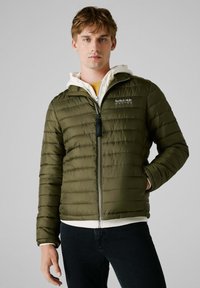 Chaqueta acolchada color verde oliva con costuras horizontales, cremallera frontal y detalle del logotipo. Presenta una sudadera blanca con capucha debajo.