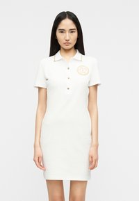 Γυναίκα με μακριά μαύρα μαλλιά, που φοράει λευκό κοντομάνικο φόρεμα πόλο με χρυσά κουμπιά και έμβλημα στο στήθος, στέκεται μπροστά σε απλό φόντο.