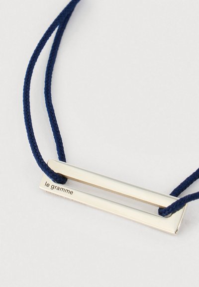 Hõbedane ristküliku kujuline ripats, millel on graveering "le gramme", tumesinise punutud nööri kaelakees, valgel taustal.