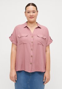 Blouse à manches courtes, à boutonnière, en tissu mauve doux, avec deux poches avant et un ourlet arrondi, ornée de boutons en bois.