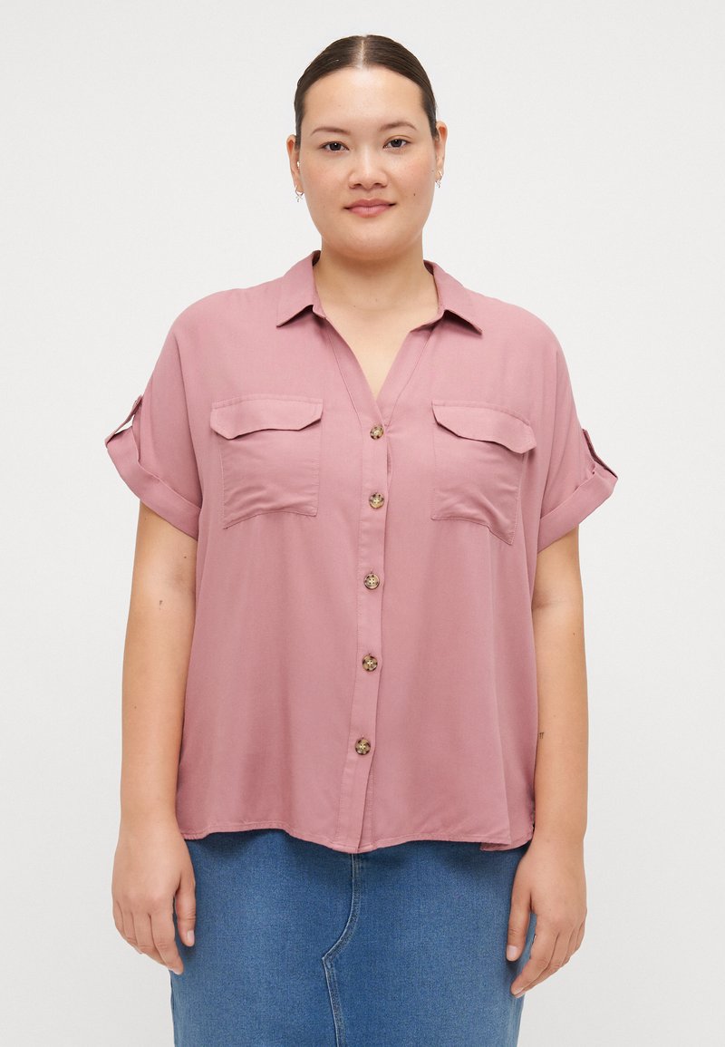 Blouse à manches courtes, à boutonnière, en tissu mauve doux, avec deux poches avant et un ourlet arrondi, ornée de boutons en bois.
