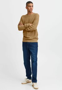 Pull en maille beige avec un col rond, associé à un jean en denim bleu foncé et des baskets blanches avec des accents beiges et marron.