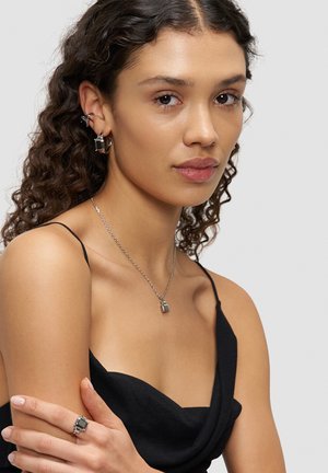 Silver necklace with a square pendant, matching square ring, and earrings; model wears a black sleeveless top with soft draping.