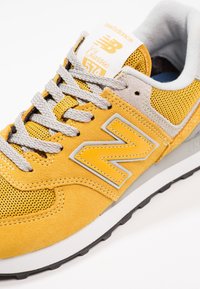 Gul New Balance 574 Classic sneaker med grå snören, överdel i mocka och mesh, samt vadderad grå krage på vit bakgrund.
