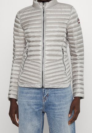 Lichtgrijze gewatteerde jas met ritssluiting aan de voorkant, met ritsen sluitbare zijzakken en een ronde logopatch op de bovenkant van de linker mouw, gedragen met een blauwe jeans.